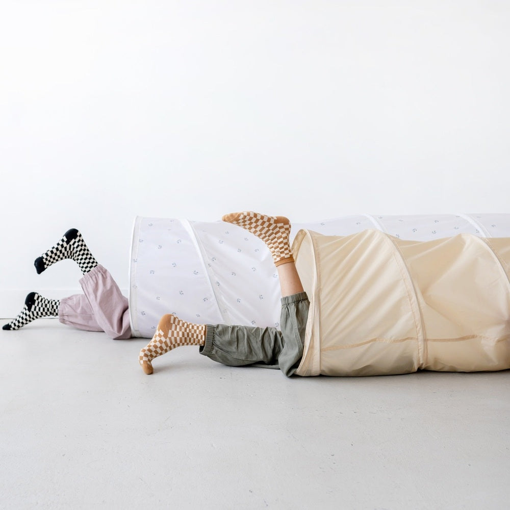 3 Sprouts Recycled Fabric Play Tunnel in beige and white; kids’ legs in checkered socks visible.