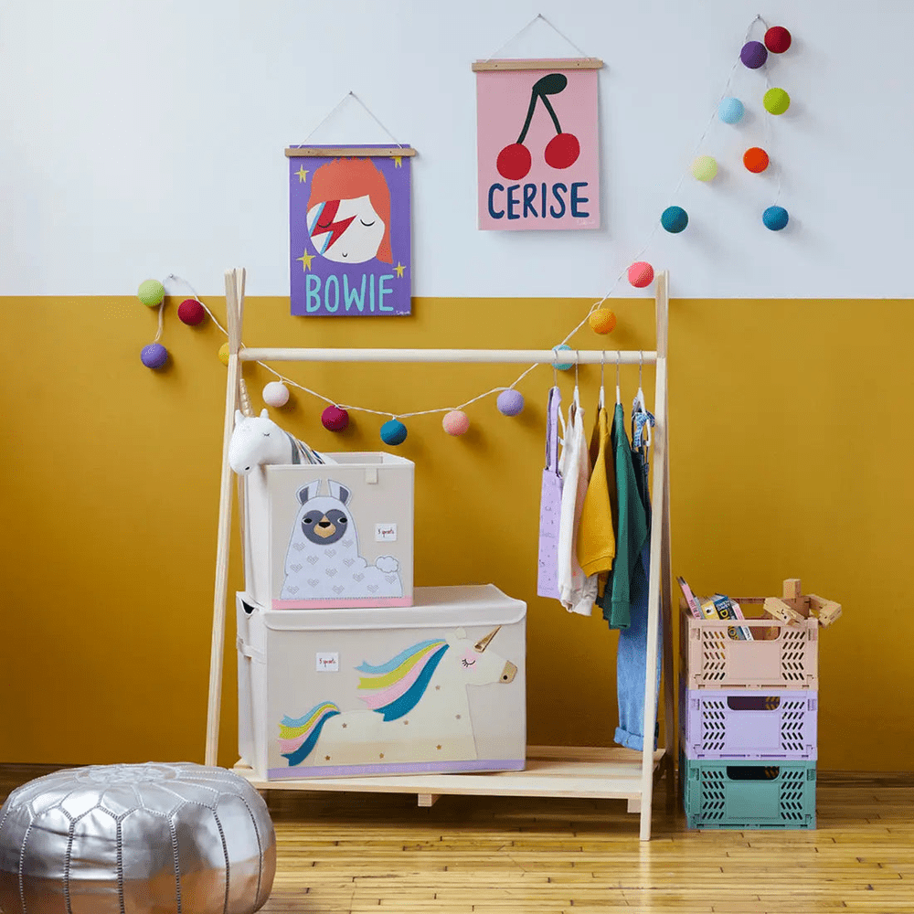 3 Sprouts Toy Chest in pastel colors with unicorn art sits under a wooden kids’ clothes rack, storing toys neatly.