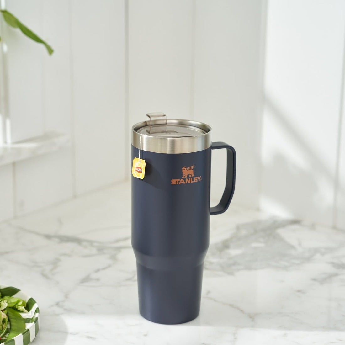 A navy blue Stanley travel mug with a stainless steel lid and handle, standing on a white marble countertop in a bright kitchen setting.