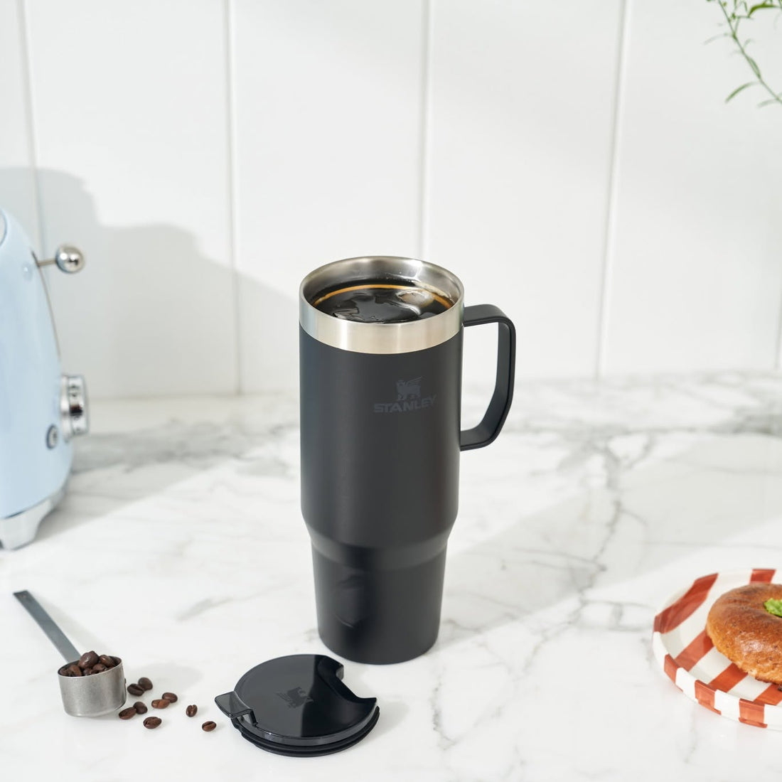 A black travel mug with a stainless steel rim and handle, shown on a marble countertop with its lid off beside coffee beans, a toaster, and a pastry.