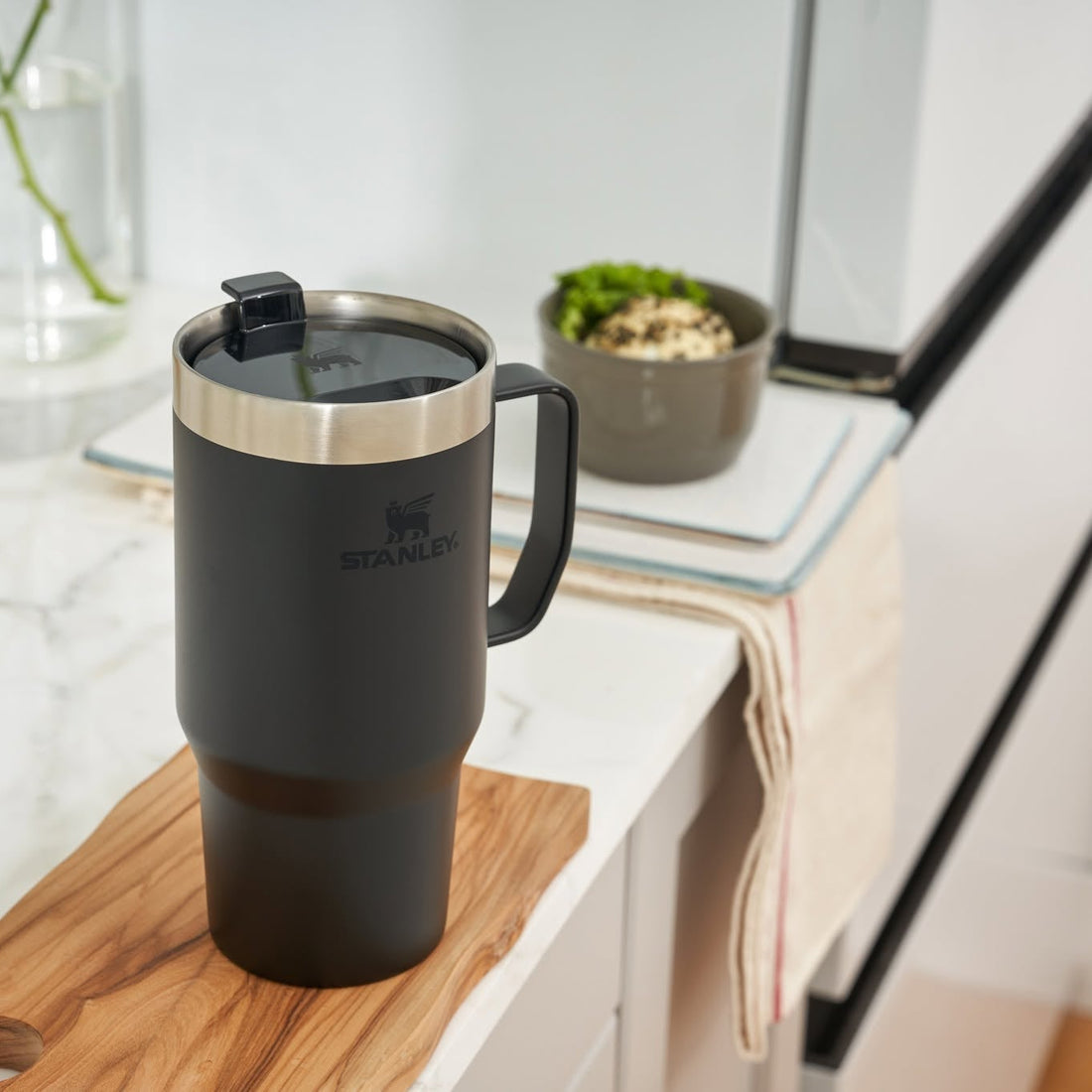 A matte black travel mug with a stainless steel rim and a black lid, resting on a wooden cutting board on a white marble countertop in a modern kitchen setting.