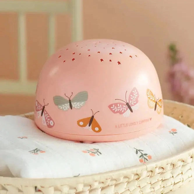A Little Lovely Company Projector Night Light in pink with butterfly decals and a starry top rests on a white cloth atop a woven basket.