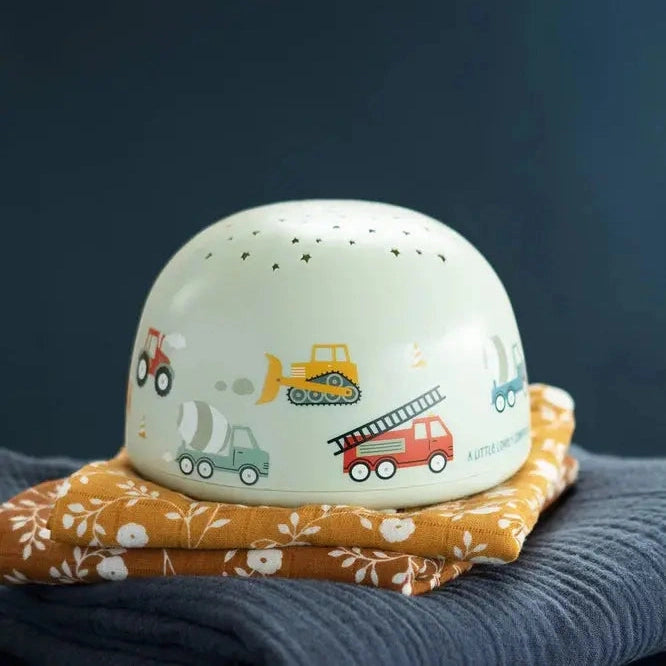 A Little Lovely Company Projector Night Light, mint ceramic with toy vehicle print, glowing softly on an orange quilt.