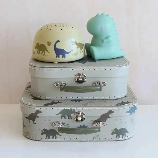 A Little Lovely Company Projector Night Light sits on stacked gray suitcases beside a pastel yellow dinosaur dome and mint lamp.