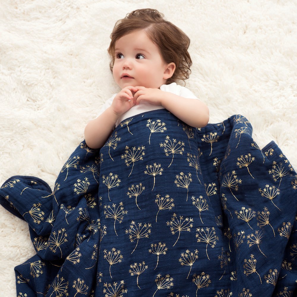 Aden+Anais Silky Soft Dream Blanket navy with metallic gold dandelion pattern enveloping a baby on a plush white rug