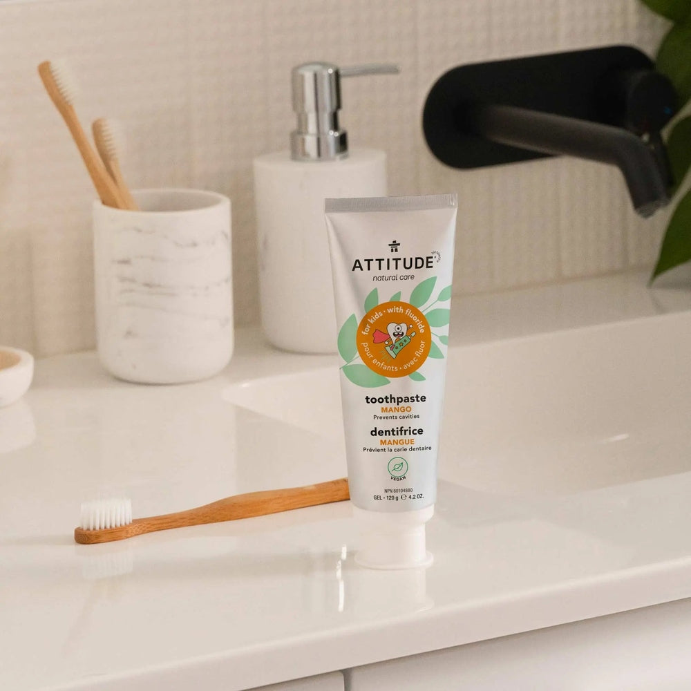 Attitude Kids Toothpaste With Fluoride Mango 120g, standing on a bathroom counter beside a wooden toothbrush.