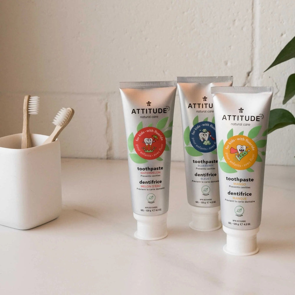ATTITUDE Kids Toothpaste With Fluoride 120G, three tubes (watermelon, blueberry, mango) standing beside toothbrushes.