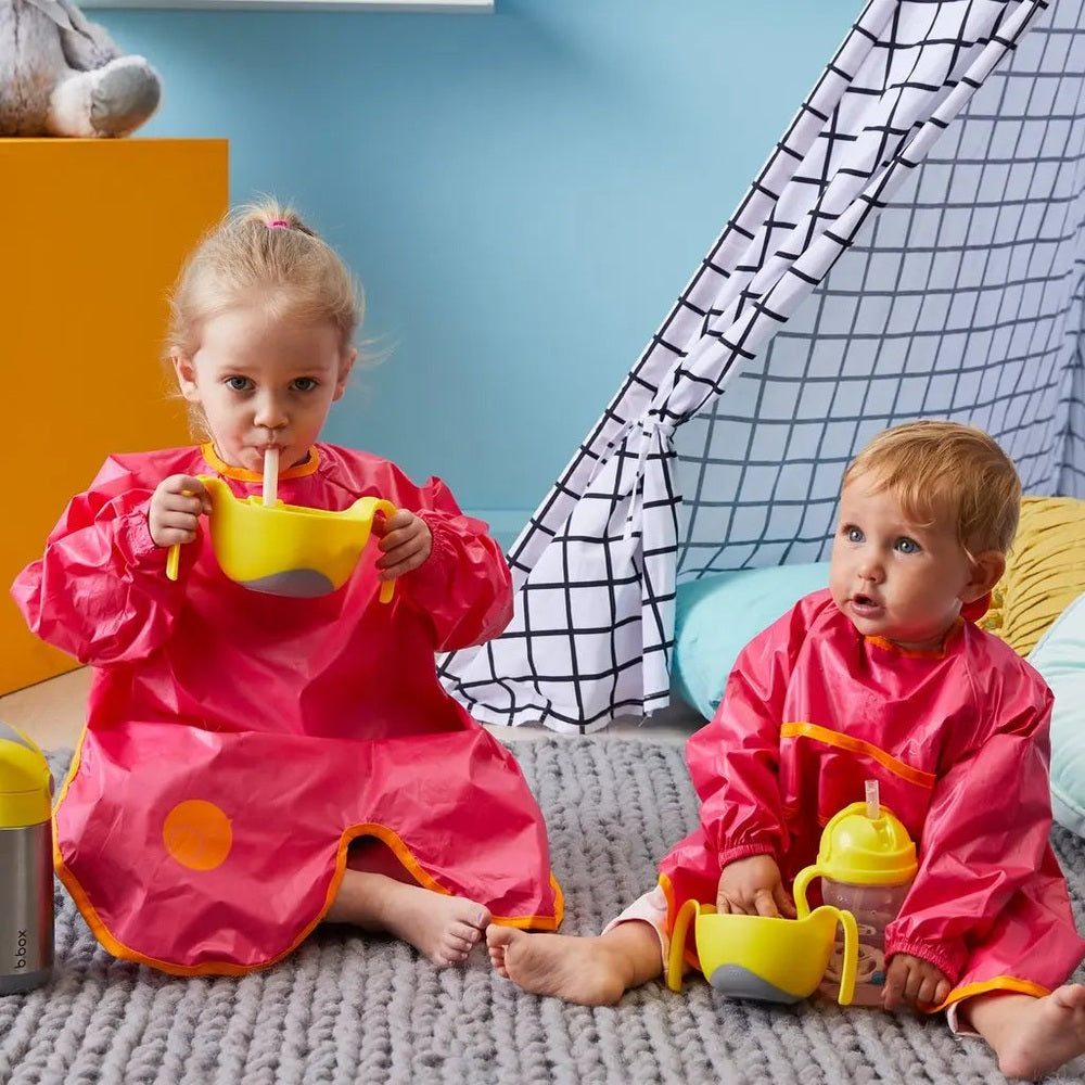 B.Box Bowl & Straw feeding set; two toddlers in pink aprons sip from bright yellow bowls with straws.