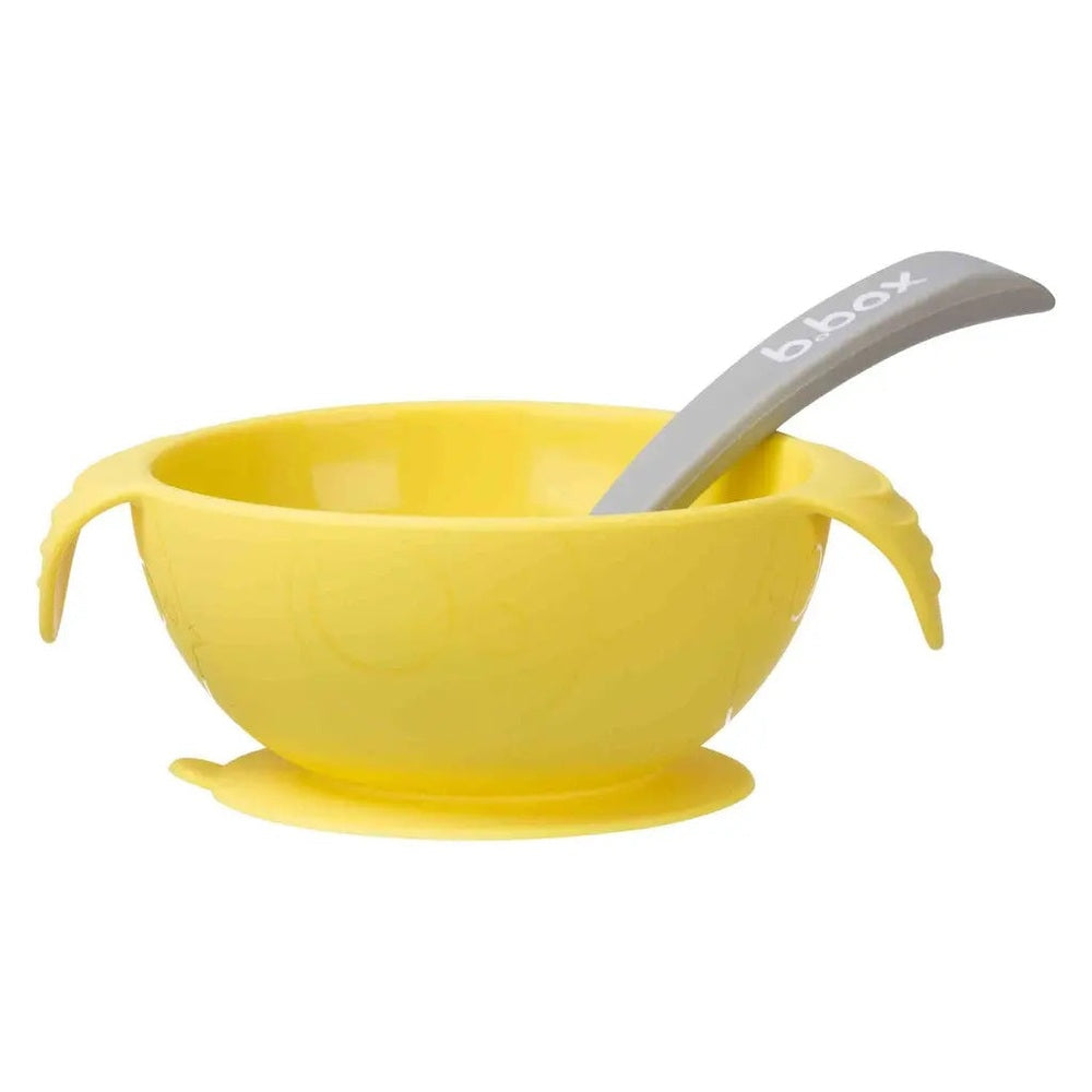 B.BOX Silicone First Feeding Set: yellow suction bowl with gray spoon resting inside to show spill-safe feeding.