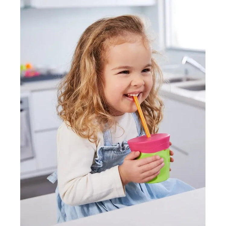 B.Box Silicone Lids Travel Pack sippy cup in green with pink lid, held by a smiling toddler sipping through an orange straw.