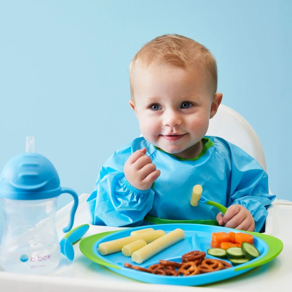 B.Box Smock Bib (6M–2Y), blue with green trim, worn by baby at mealtime catching spills.