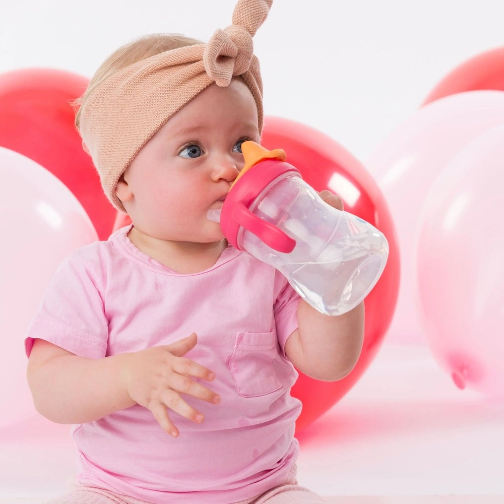 B.Box Transition Value Pack sippy cup with pink lid, held by a baby sipping through the orange spout.