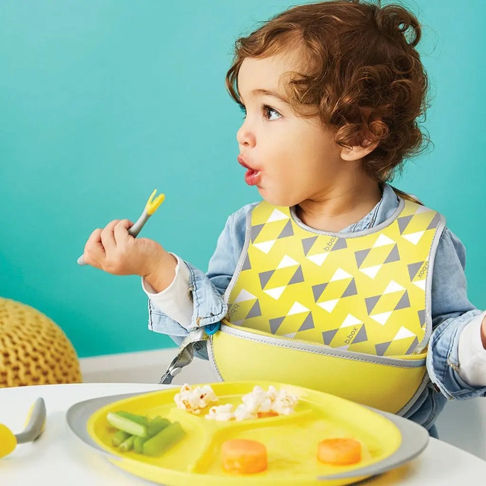 B.Box Travel Bib in bright yellow with grey triangles; toddler happily eats with a spoon, showing easy-meal coverage.
