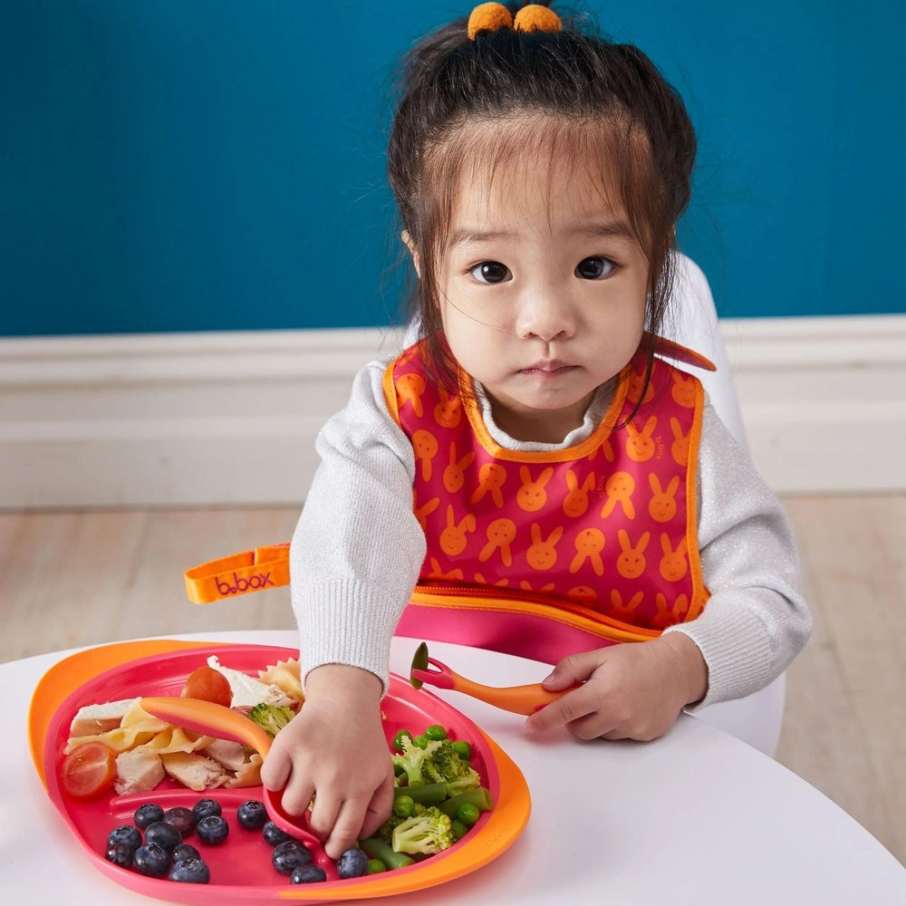 B.Box Travel Bib in orange bunny pattern; toddler reaches for blueberries on a colorful plate.