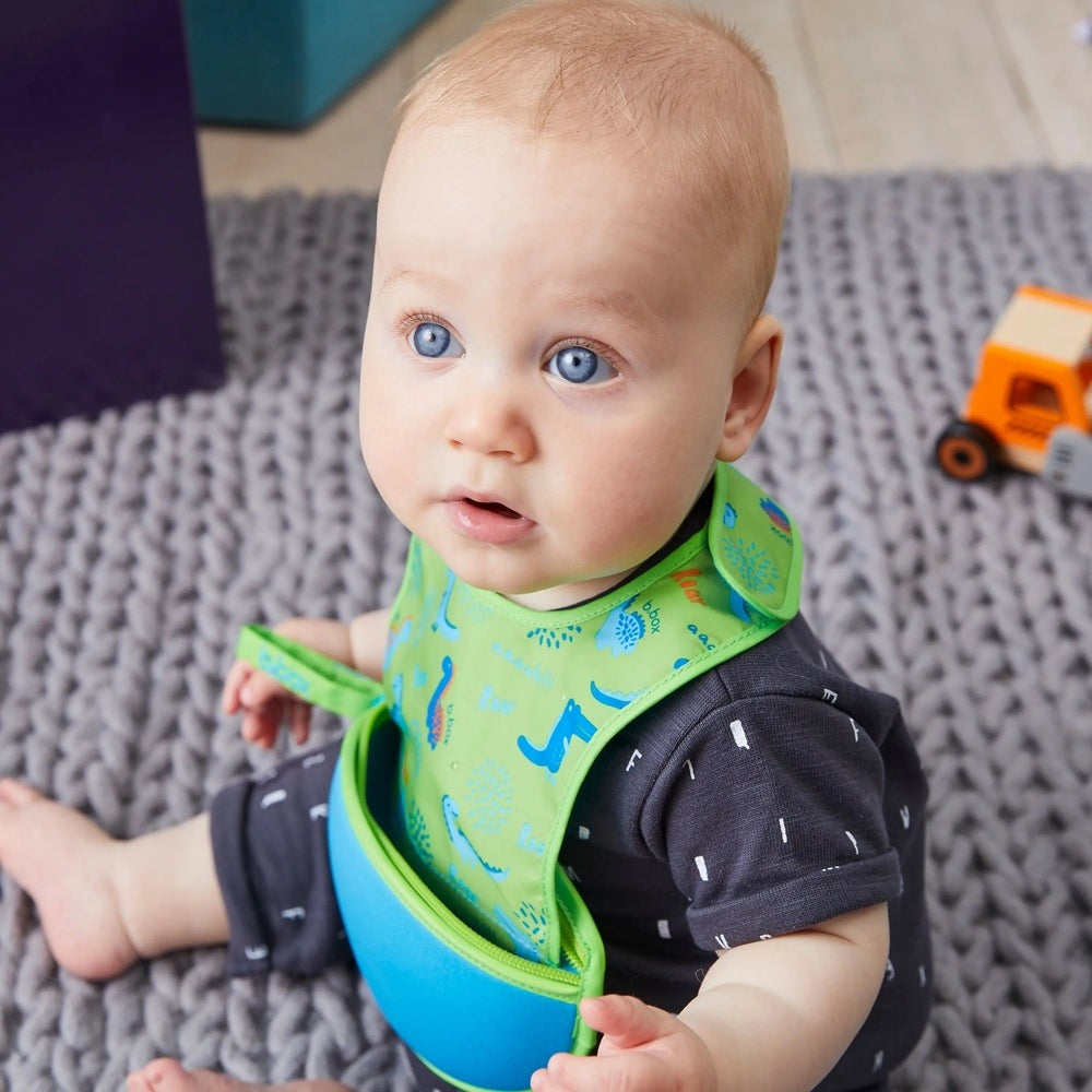 B.Box Travel Bib in bright green with a blue pocket, worn by a curious baby sitting on a soft gray rug.