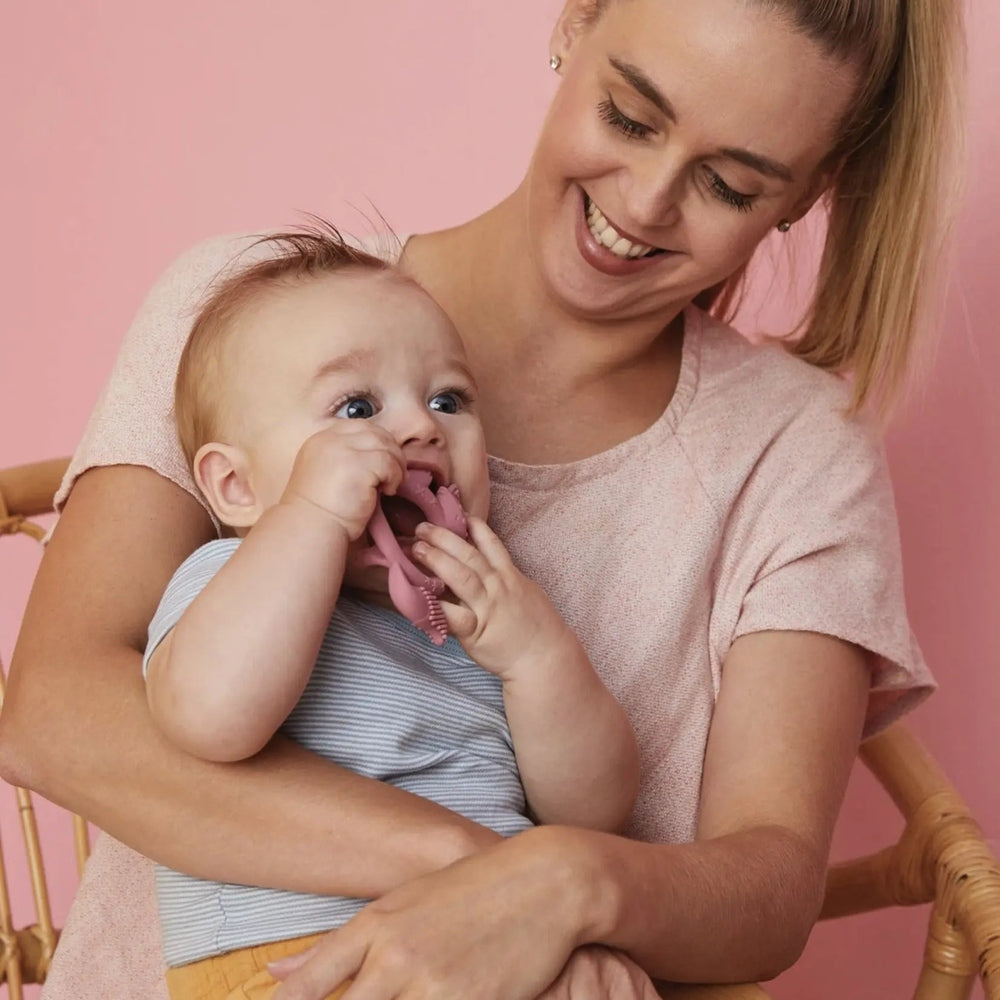 B.Box Wrist Teether pink silicone worn on baby's wrist as mom cuddles and baby chews happily.