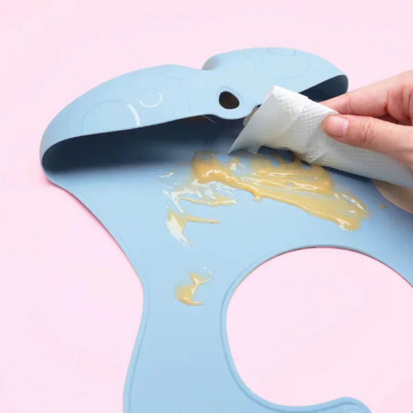 B.box silicone bib in light blue with open-neck design being wiped with a napkin.