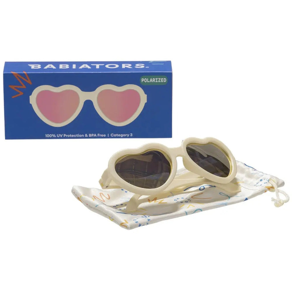 Babiators Polarized Mirrored Sunglasses Heart Shape Sweet Cream resting on a patterned drawstring pouch with pink lenses.