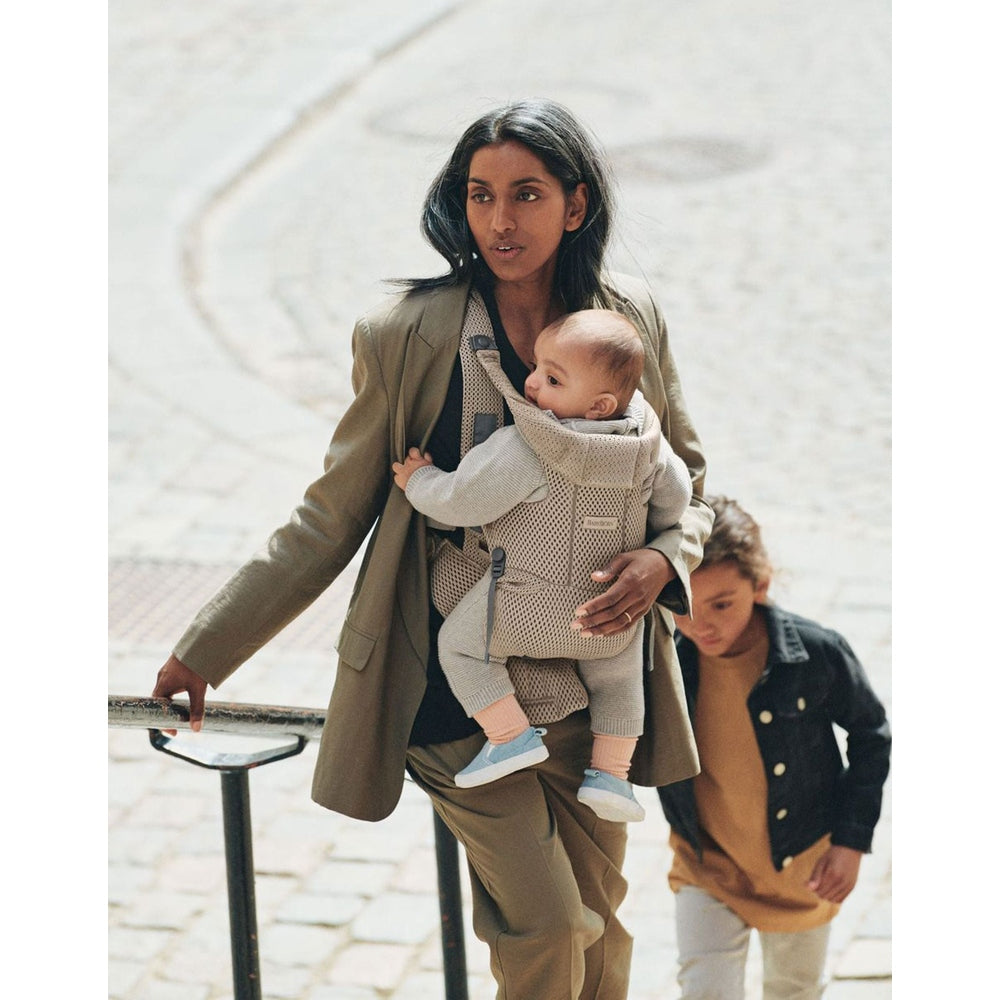 Baby Björn Baby Carrier Free beige worn by a mother with the baby strapped to her chest on outdoor stairs.