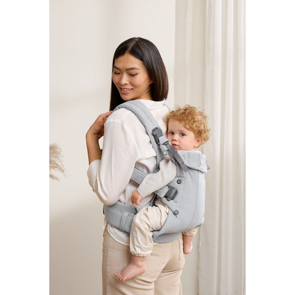 BabyBjörn Baby Carrier Harmony in light gray worn by a smiling mom with toddler securely on her back.