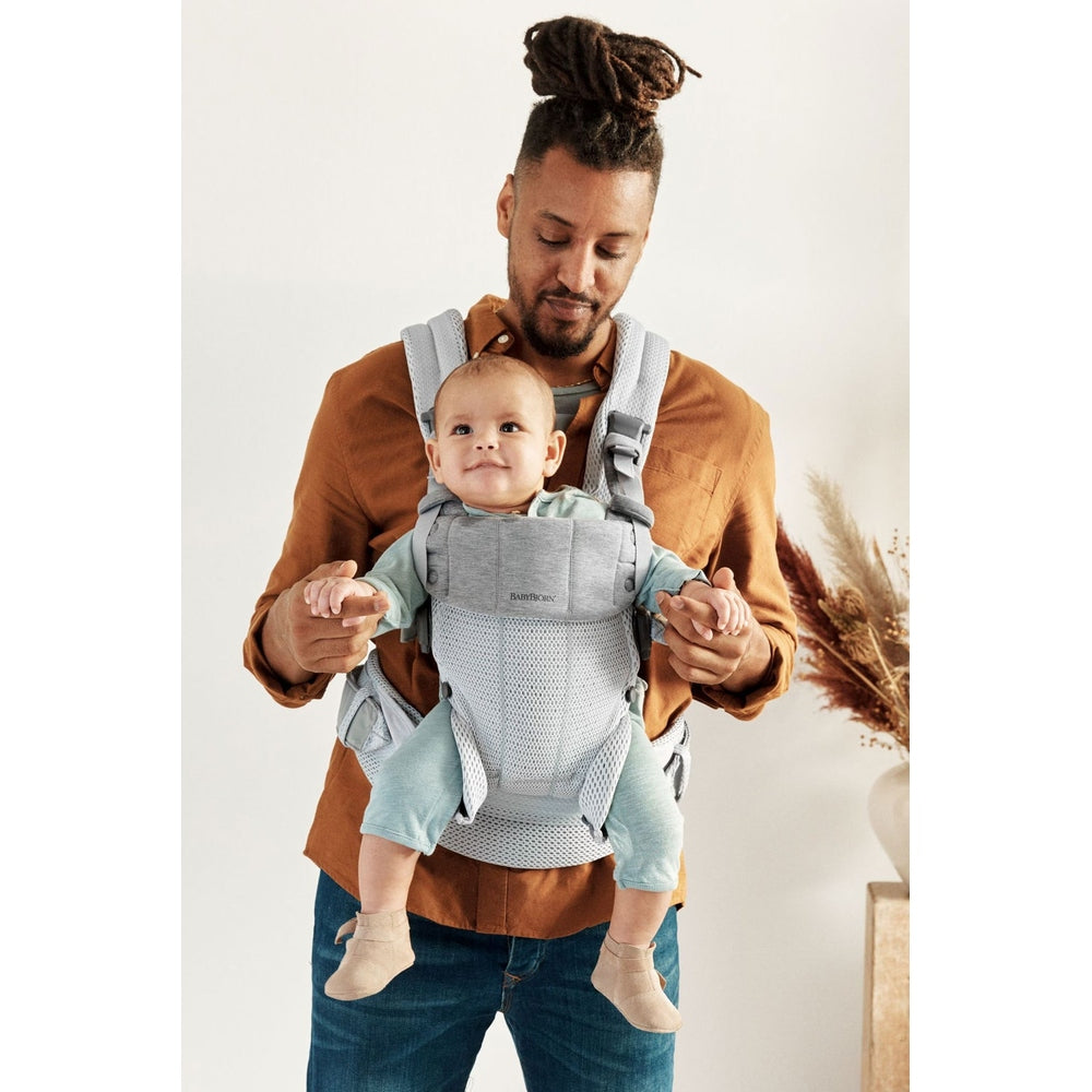Father wearing BabyBjörn Baby Carrier Harmony in light gray with breathable mesh straps, carrying smiling baby.