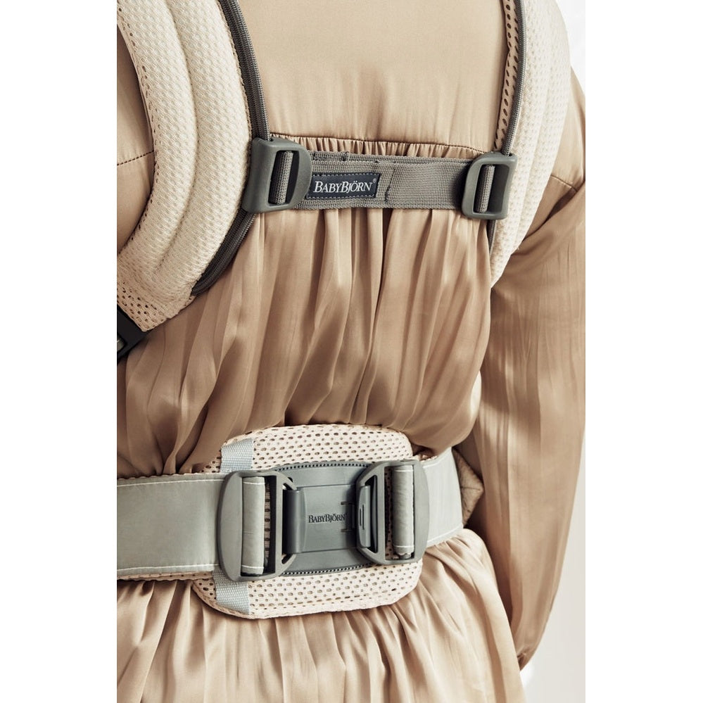 BabyBjörn Harmony Baby Carrier worn on a beige outfit; gray padded straps and buckle-secured waist belt.