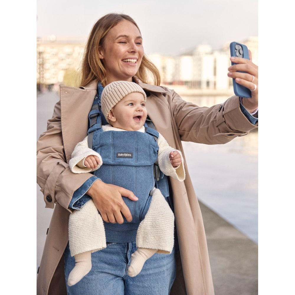 Mother wears BABY BJÖRN BabyBjörn Harmony baby carrier, smiling baby secured in denim, taking a selfie by the water.