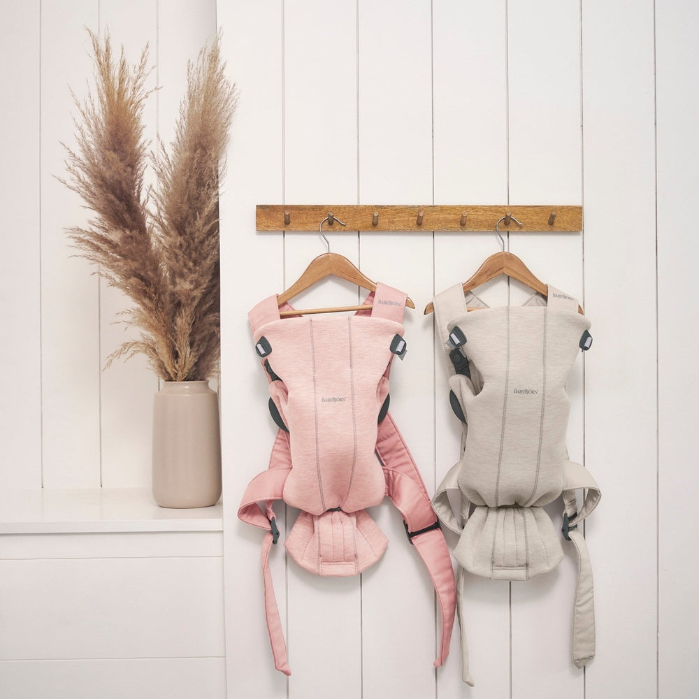 BabyBjörn Baby Carrier Mini in pink and gray, two carriers hung on a wooden rack beside a pampas grass vase.