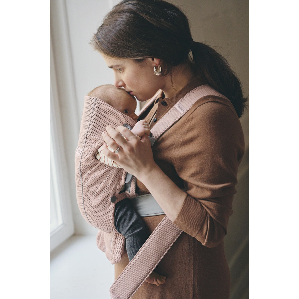 BabyBjörn Baby Carrier Mini in soft pink; mom cradles baby close with cushioned mesh straps.