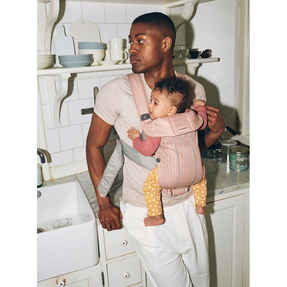 Father wearing BabyBjörn Baby Carrier Mini in soft pink, carrying baby close with padded straps in a cozy kitchen.