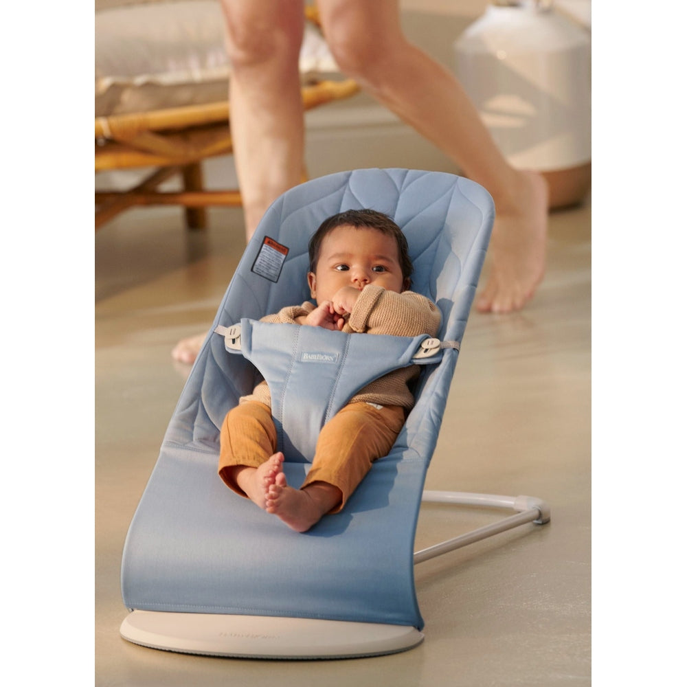 BabyBjörn Bouncer Bliss in light blue, baby secured in reclined seat with harness as an adult walks nearby.