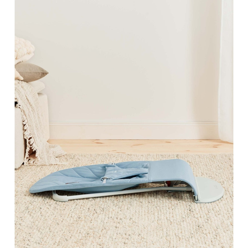 BabyBjörn Bouncer Bliss in blue, folded compactly on a beige rug to show its space-saving design.