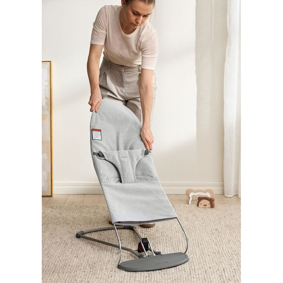 Mother adjusts BabyBjörn Bouncer Bliss in gray fabric, highlighting sturdy metal frame and compact base in a serene room.
