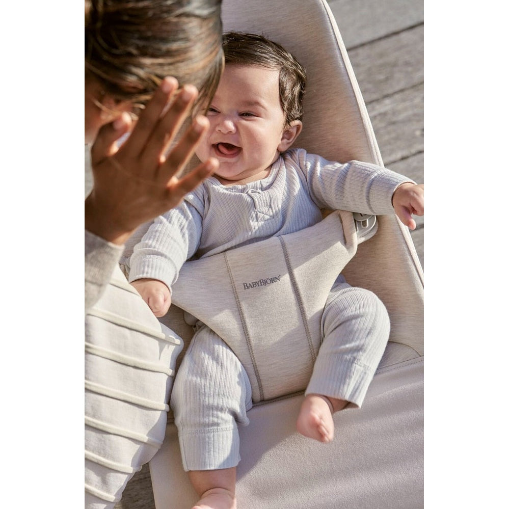 BabyBjörn Bouncer Bliss in beige fabric; smiling baby secured in harness while caregiver plays nearby outdoors.