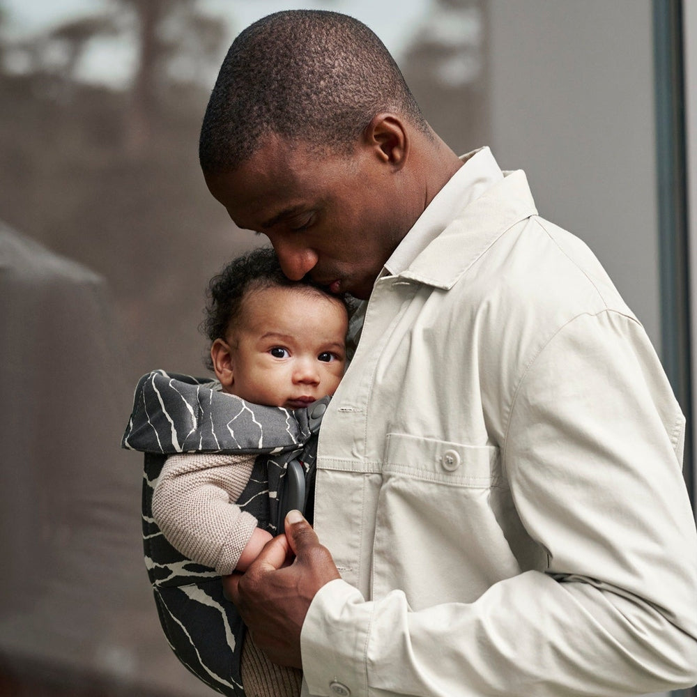 Man holds a baby in a BabyBjörn Bouncer Bliss Black Mesh Bundle with Baby Carrier Mini Landscape Holiday Bundle, demonstrating front-carry comfort.