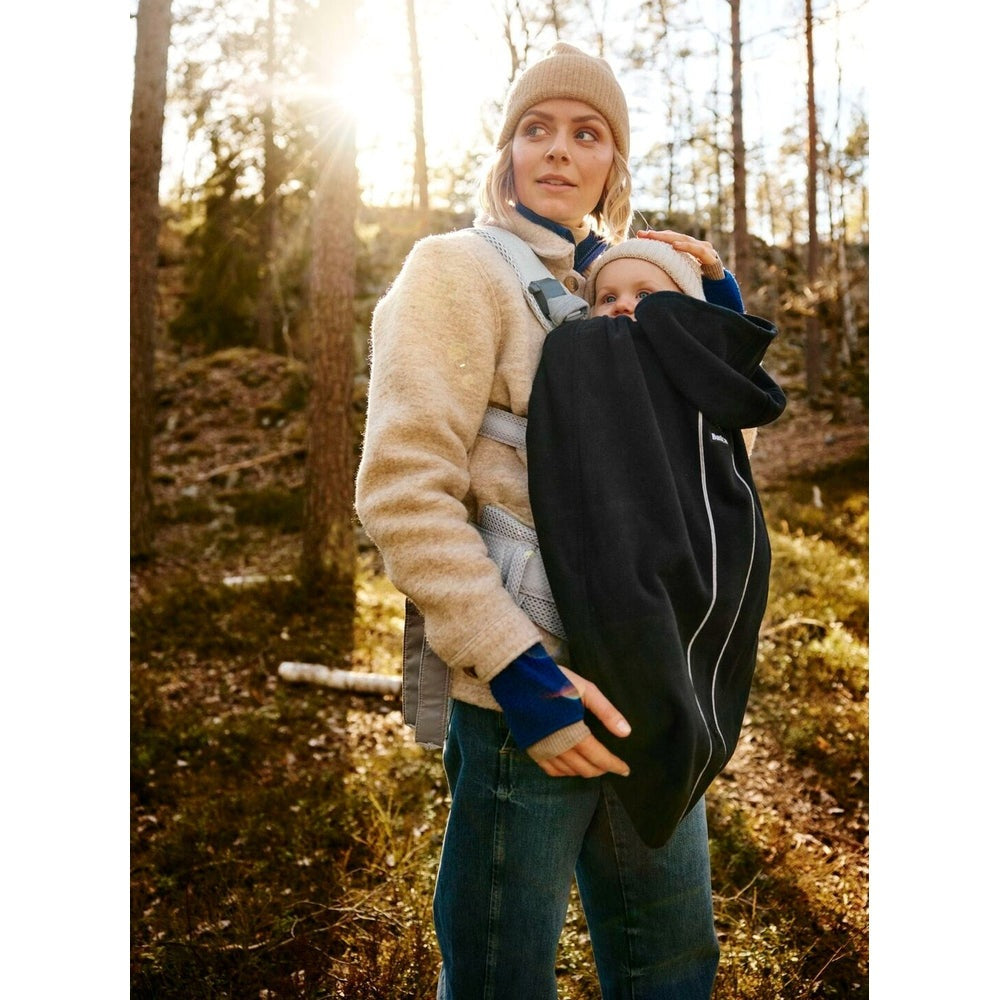 BabyBjörn Cover for Baby Carrier in black shields baby as parent walks through sunlit forest.