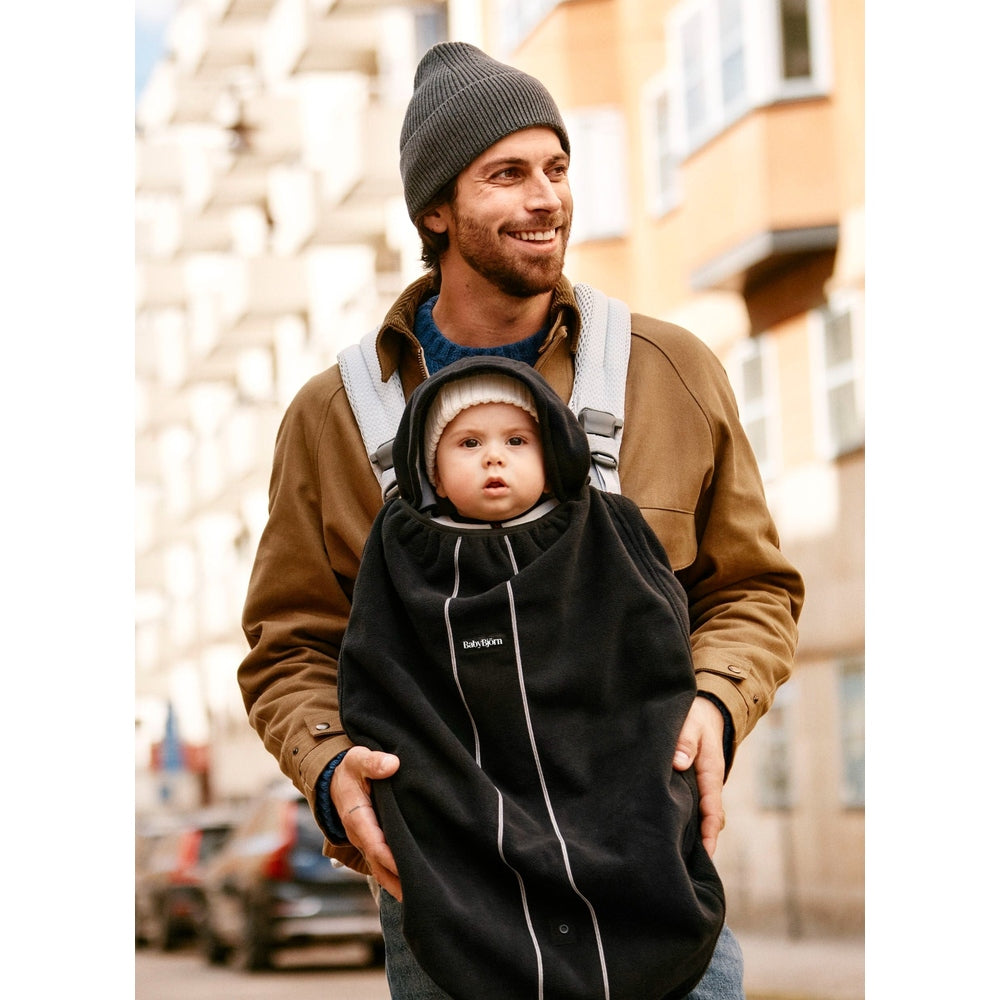 BabyBjörn Cover for Baby Carrier in black with white seams, worn by a man outdoors carrying a baby.