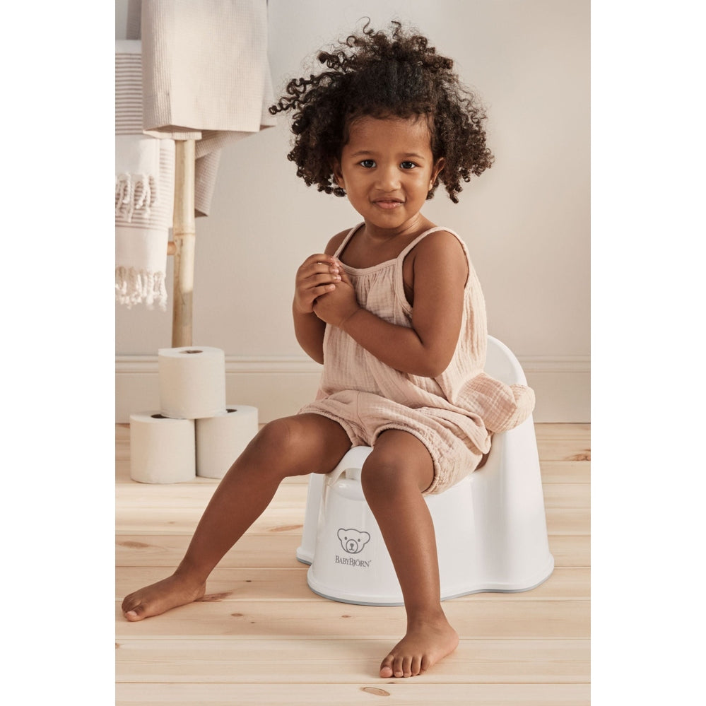 BabyBjörn Potty Chair, white with logo; toddler girl in beige outfit sits and smiles.
