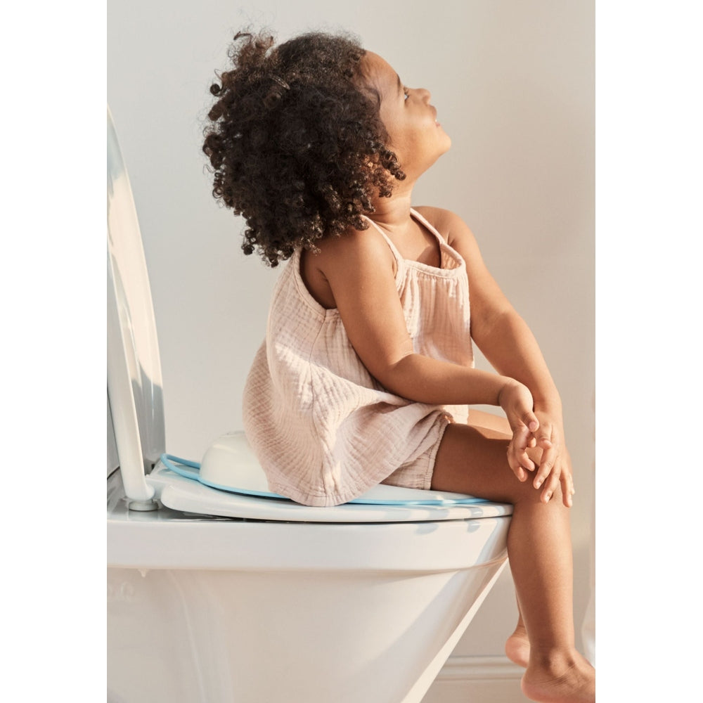 BabyBjörn Toilet Trainer on a bathroom toilet; a toddler in a pink romper sits on the blue trainer ring.
