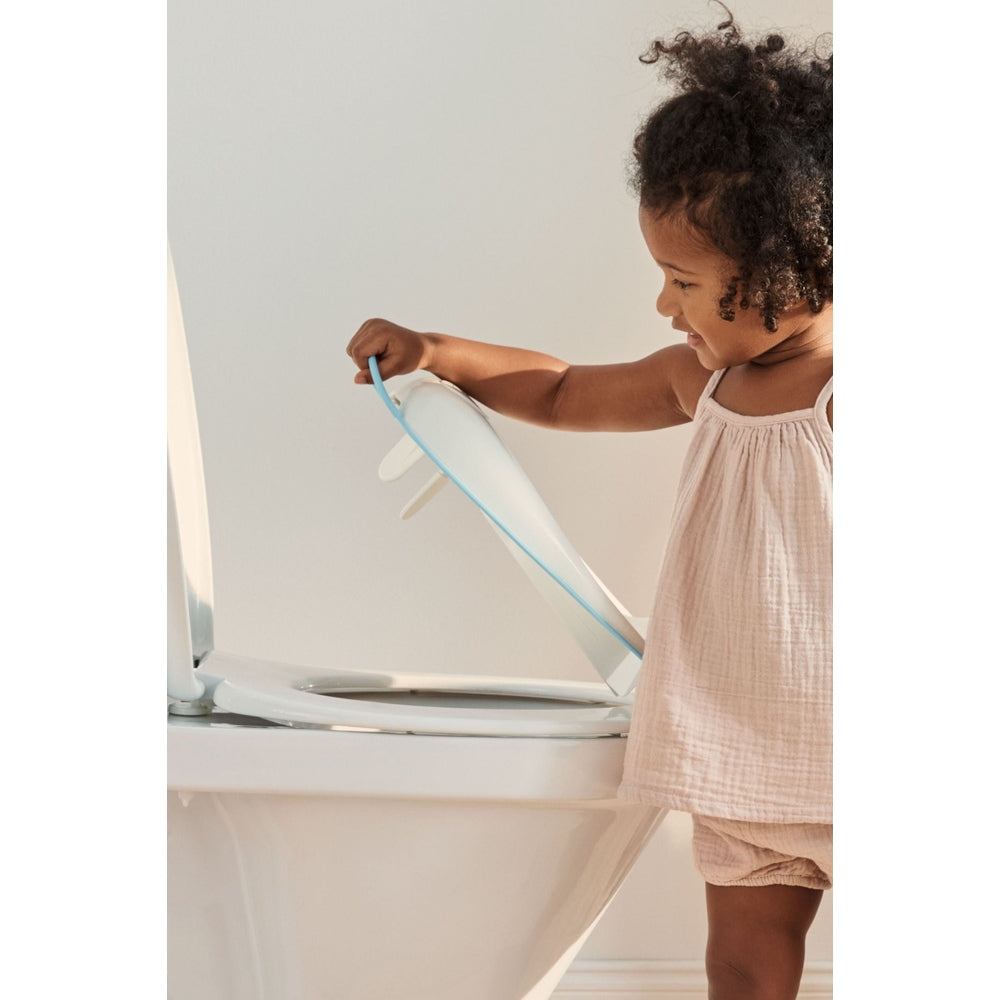BabyBjörn Toilet Trainer in pale blue, toddler lifting the training seat onto a white toilet.