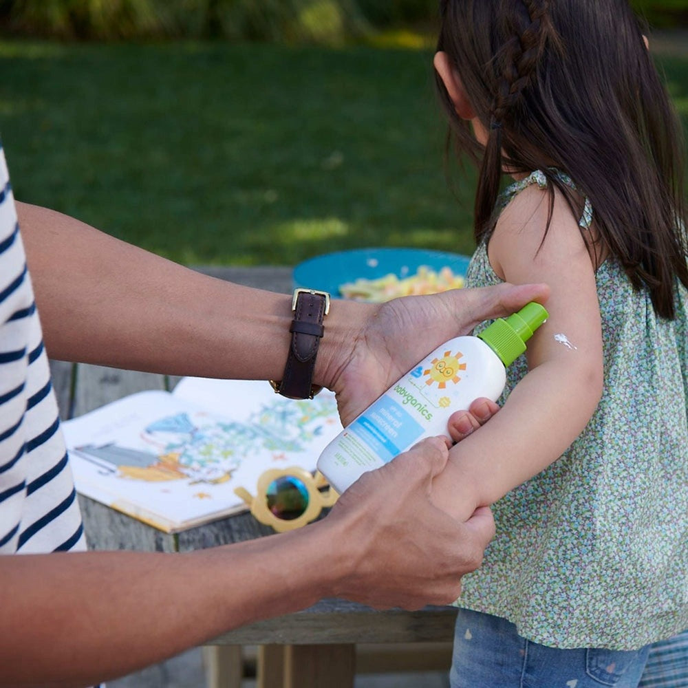 Babyganics Mineral Sunscreen Spray SPF50 bottle with green cap applied to a child's arm outdoors