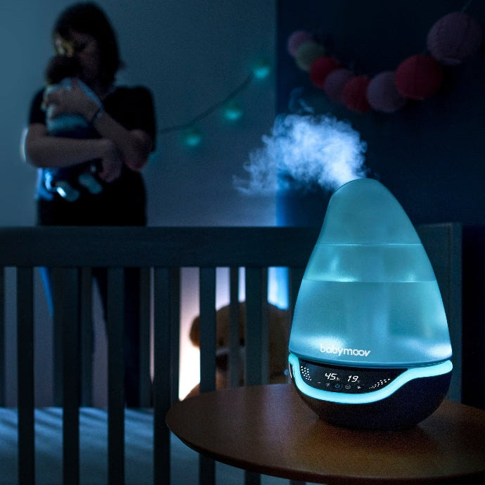 Babymoov Hygro Plus 3-In-1 Baby Humidifier with blue glow releasing mist on a wooden table beside a crib.