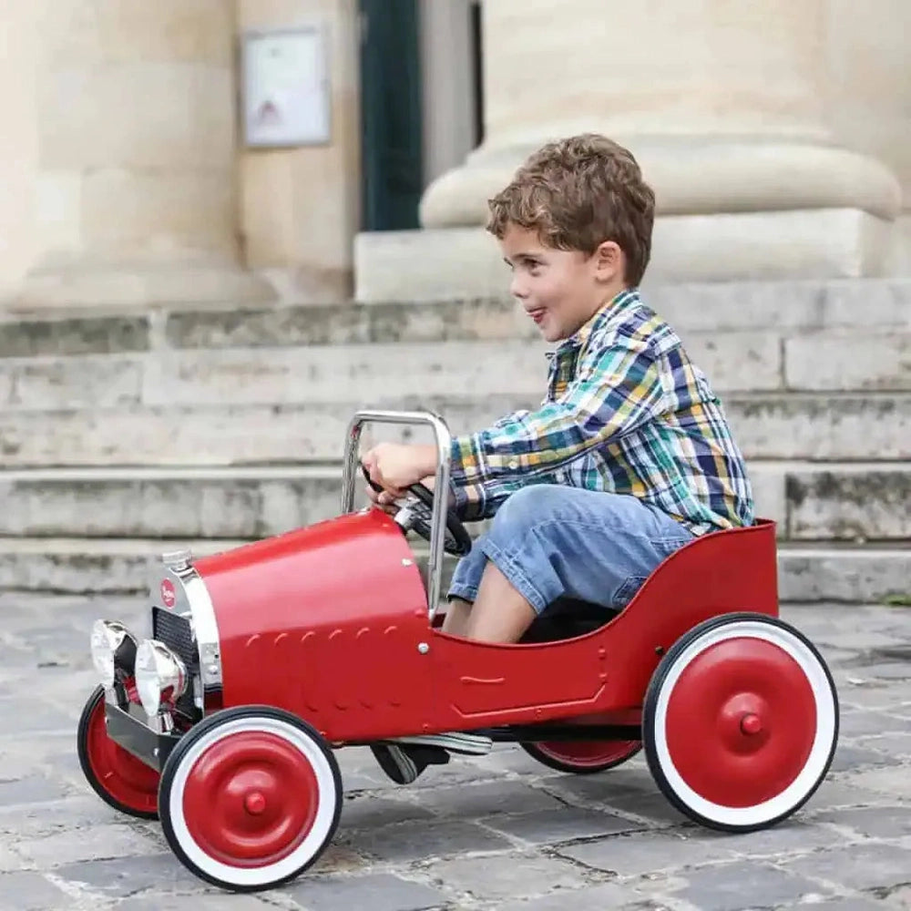 Baghera Ride-On Classic Pedal Car red with white-wall wheels, boy riding on cobblestones.