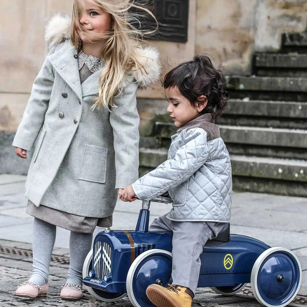 Two kids outdoors with a blue Baghera Ride-On Speedster Citroën La Petite Rosalie; one child rides while the other holds hands.