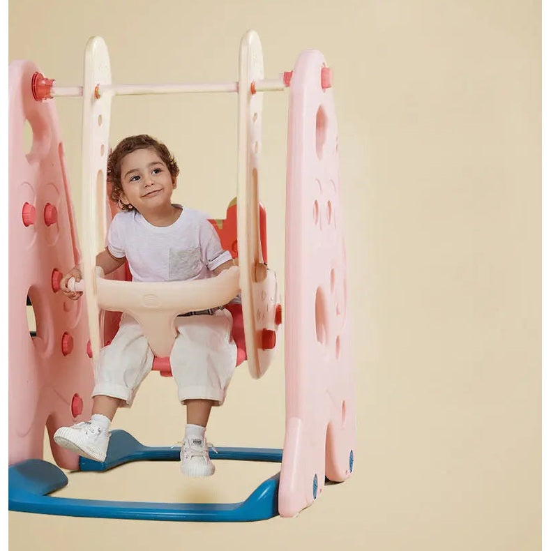 Bc Babycare 5 In 1 Slide playground swing set, pink frame with blue base, child seated and gripping the bar.