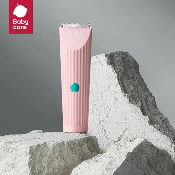 BC Babycare Baby Hair Clipper in pink with teal button, standing upright on sandy rocks beside a white mound