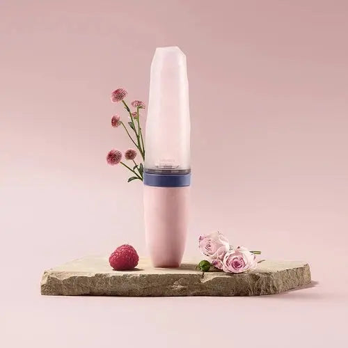 BC Babycare Feeding Spoon Dispenser standing upright in pink with a blue band, on a stone slab beside flowers.