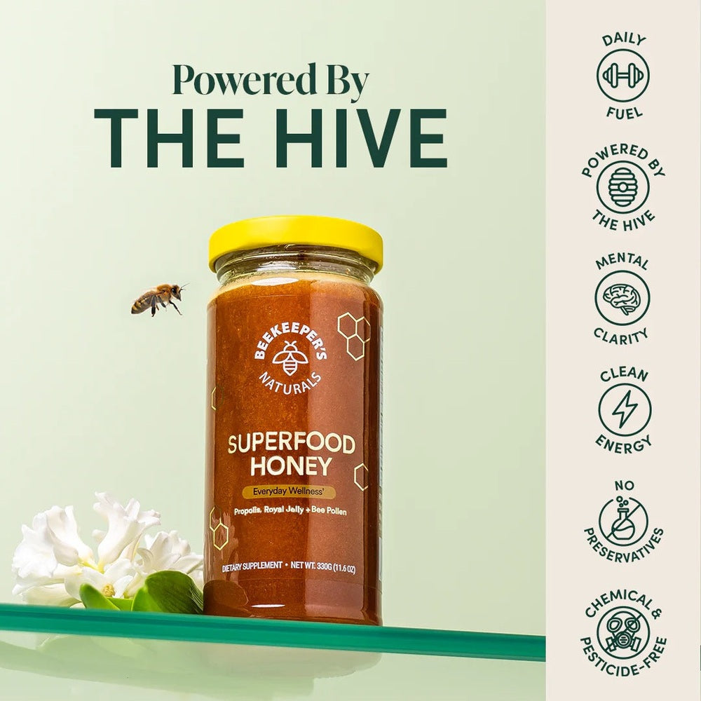 Beekeeper's Naturals Superfood Honey 330g in an amber jar with a bright yellow lid, beside a bee and white flowers.