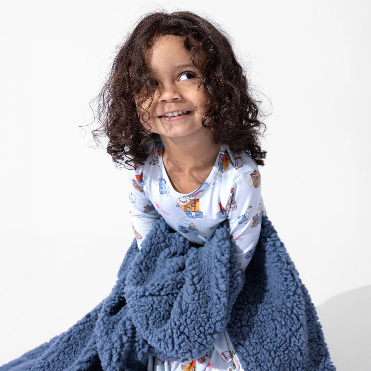 Bellabu Bear Bamboo Kids Pajamas Vintage Suitcases; child sits smiling, wrapped in plush blue fleece blanket with suitcase prints.