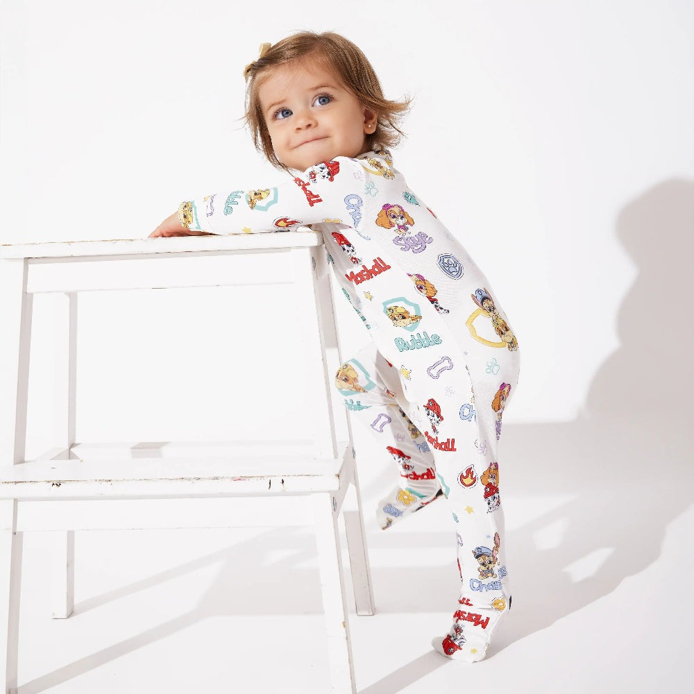 Bellabu Bear Playful Pups Bamboo Convertible Footie Paw Patrol: toddler leans on a white stool in printed sleepwear.
